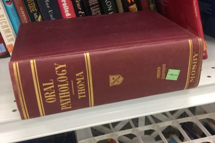 Books sit on a shelf at a thrift store, and a large brown book sits in front of them, titled Oral Pathology, author's last name is Thoma.