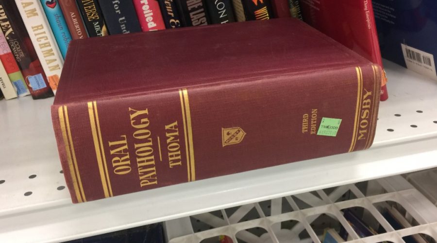 Books sit on a shelf at a thrift store, and a large brown book sits in front of them, titled Oral Pathology, author's last name is Thoma.