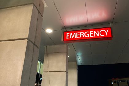 Photo of the entrance to a hospital emergency room, or Accident and Emergency department for those in the U.K. A lighted sign that says "Emergency" hangs from a gray tiled ceiling above the entrance.