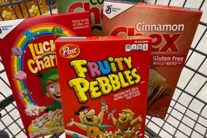 Boxes of cereal sit in a shopping cart. Clockwise from left: Lucky Charms, Corn Chex, Cinnamon Chex, and Fruity Pebbles.