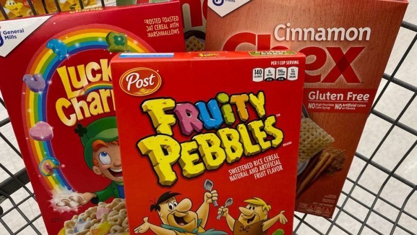 Boxes of cereal sit in a shopping cart. Clockwise from left: Lucky Charms, Corn Chex, Cinnamon Chex, and Fruity Pebbles.