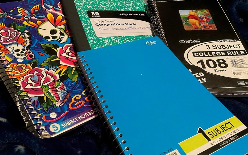 A group of notebooks lays on a bed with a dark blue blanket, starting with a five-subject Ed Hardy-themed Lisa Frank notebook, a green composition notebook, a small three-subject notebook with a black cover and a Winnie the Pooh sticker on the front of it, and a small plain light blue notebook in the foreground. Used for illustrative purposes for Write Down Your Story Day.