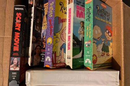 An illustrative photo depicting an open cardboard box containing copies of old VHS tapes including Scary Movie, Labyrinth, Doom Runners, Rugrats Tommy Troubles, Drowning Mona, Rugrats Tales from the Crib, and a videotape in a plastic white case without a label for VCR Day 2021.