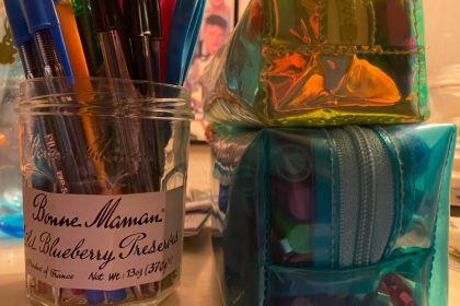 An old jelly jar repurposed to serve as a place to store unused ballpoint pens in red, blue, and black, with an orange pen in the foreground, and a green one off to the left. Two rectangular pencil cases sit nearby, the bottom one in clear blue and full of pens, markers, and a mechanical pencil. Under it is a clear neon green case lined in white, which has highlighters, more pens, and an eraser inside it for National Ballpoint Pen Day 2021