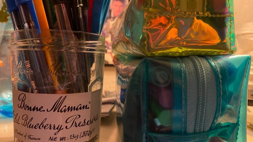 An old jelly jar repurposed to serve as a place to store unused ballpoint pens in red, blue, and black, with an orange pen in the foreground, and a green one off to the left. Two rectangular pencil cases sit nearby, the bottom one in clear blue and full of pens, markers, and a mechanical pencil. Under it is a clear neon green case lined in white, which has highlighters, more pens, and an eraser inside it for National Ballpoint Pen Day 2021