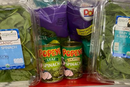 Two bags of Dole branded fresh spinach are in the back of a shopping cart seat, with one plastic container of Josie's Organics branded spinach on the far left and right sides. A can of Popeye branded leaf spinach and a can of chopped spinach are in front of the Dole bags, next to each other.