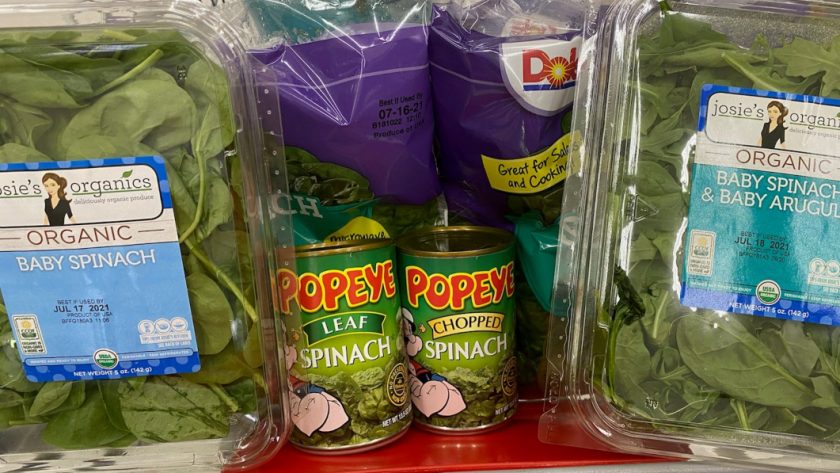 Two bags of Dole branded fresh spinach are in the back of a shopping cart seat, with one plastic container of Josie's Organics branded spinach on the far left and right sides. A can of Popeye branded leaf spinach and a can of chopped spinach are in front of the Dole bags, next to each other.