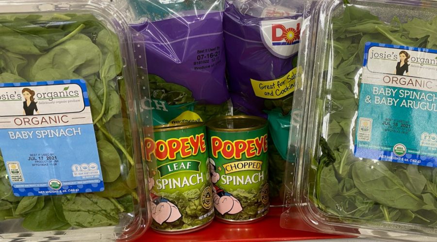 Two bags of Dole branded fresh spinach are in the back of a shopping cart seat, with one plastic container of Josie's Organics branded spinach on the far left and right sides. A can of Popeye branded leaf spinach and a can of chopped spinach are in front of the Dole bags, next to each other.