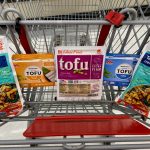Packages of tofu sit in a grocery cart seat, with a package of tempeh on the far left and right sides.