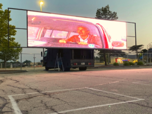 A pic of a large drive-in movie screen showing a scene from Field of Dreams, released in 1989, pic taken in the evening just toward the end of sunset during the summer of 2022. The screen sits on a disused parking lot, and the pic was taken while I was sitting on the ground on an outdoor blanket.