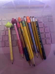 A bundle of pencils, shortened from use, sits in an old pink Easter bucket, which sits on top of a laptop computer. Some of the pencils have cap erasers. Some are glitter pencils in yellow, pink, and purple. Others are basic yellow pencils, and some are promotional pencils that were freebies from the fair. One is a My First Ticonderoga.