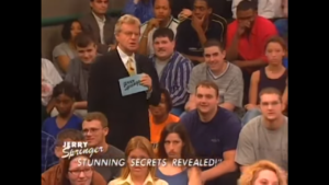 A screenshot of an episode of the Jerry Springer Show from 1999, where Jerry stands among the seated audience wearing a suit and tie, holding a microphone, and cue cards with the Jerry Springer Show logo on the backs.