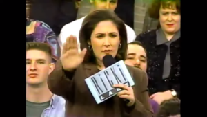 A screenshot from an episode of Ricki Lake on YouTube from the 1990s, with Ricki wearing a brown suit, holding a microphone, and cue cards with the Ricki Lake logo on the back. She stands among seated audience members. 