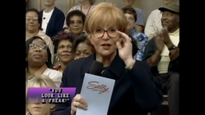 A screenshot from an old episode of the Sally Jessy Raphael show from the 1990s, uploaded onto YouTube. Sally stands among the seated audience, wearing red glasses, a black suit, and holding cue cards w/ the Sally logo on the backs. 