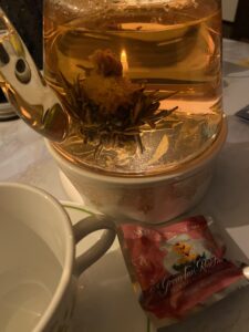 A close up of the Teabloom Buckingham Palace teapot set, filled with hot water, the included tealight candle lit and placed in the warmer, and a tea blooming flower sits at the bottom of the pot. In the foreground is a vintage Pyrex teacup and matching saucer, and off to the side is the unopened blooming tea ball in My Growing Love. 