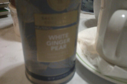A photo of a canister of White Ginger Pear tea from Teabloom, sitting on a white marble-hued coffee table, next to an old white mug full of tea.