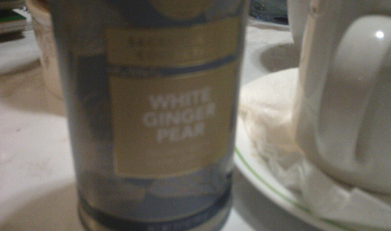 A photo of a canister of White Ginger Pear tea from Teabloom, sitting on a white marble-hued coffee table, next to an old white mug full of tea.