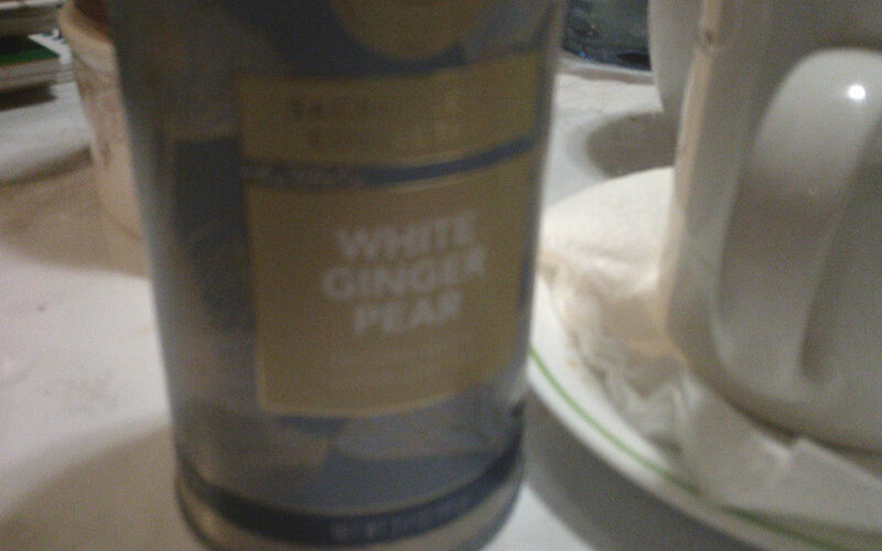 A photo of a canister of White Ginger Pear tea from Teabloom, sitting on a white marble-hued coffee table, next to an old white mug full of tea.