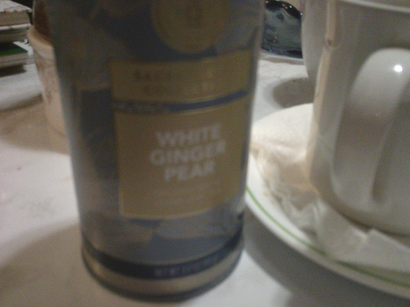 A photo of a canister of White Ginger Pear tea from Teabloom, sitting on a white marble-hued coffee table, next to an old white mug full of tea.